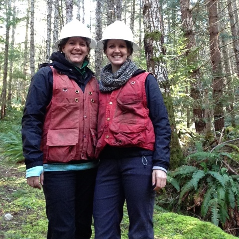 Two women smiling the the camera while wearing white hard hats and bright red vests. They are standing in a forest.